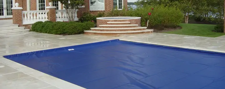 A rectangular swimming pool is covered with a blue safety cover on a stone patio near a brick building. There are steps leading up to a circular hot tub and landscaped greenery in the background.
