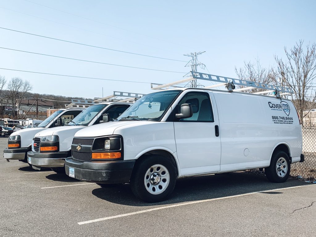 Three white work vans with ladders on top are parked in a row in a lot. The vans have company logos and contact information on the side. Trees, power lines, and buildings are visible in the background.