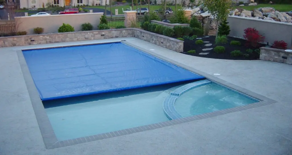 A backyard pool with a blue safety cover partially closed, next to a round hot tub. The pool area is surrounded by landscaping, stone walls, and shrubs.
