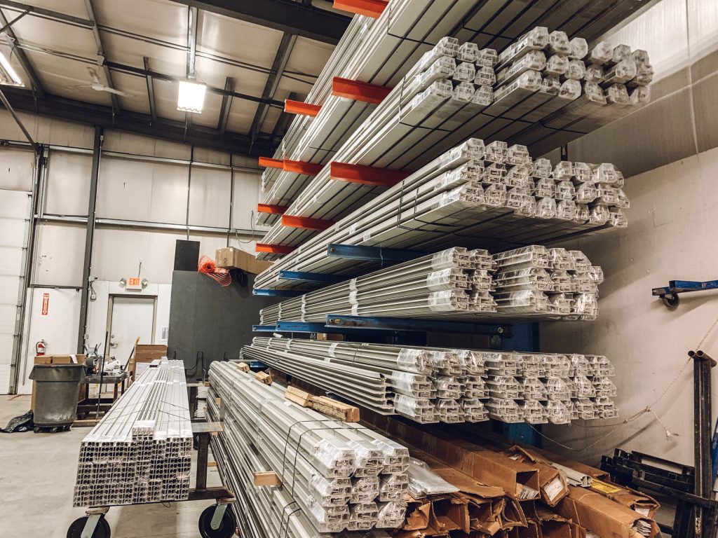 Metal bars and rectangular aluminum profiles are stacked on red and blue industrial shelving inside a warehouse with high ceilings and fluorescent lighting. The organized materials are ready for use or shipment.