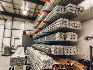 Metal bars and rectangular aluminum profiles are stacked on red and blue industrial shelving inside a warehouse with high ceilings and fluorescent lighting. The organized materials are ready for use or shipment.