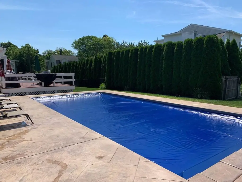 A backyard swimming pool covered with a blue pool cover, surrounded by a concrete patio, with lounge chairs nearby and tall green hedges lining the fence. A deck and house are visible in the background under a clear sky.