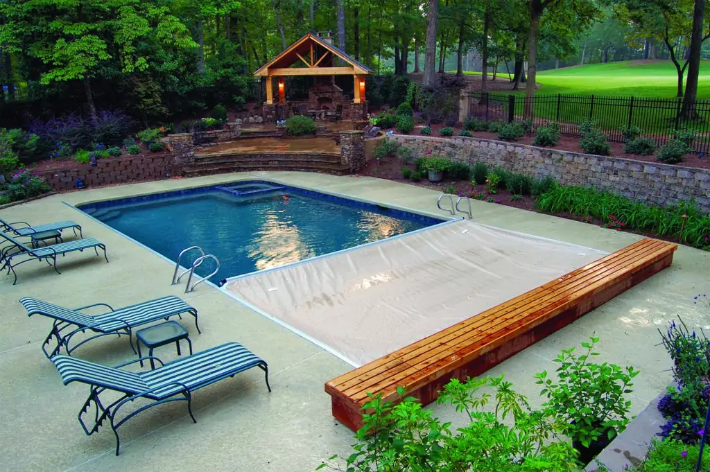 A backyard pool with a partially extended pool cover, surrounded by lounge chairs and landscaping, with a wooden pergola and trees in the background.