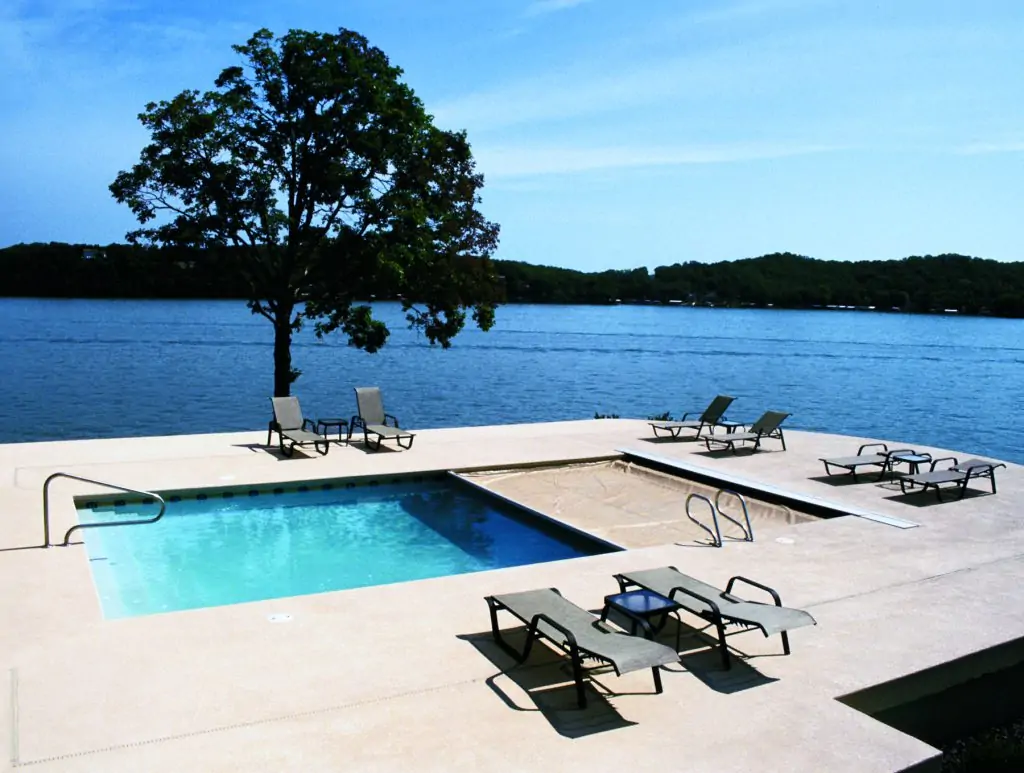 A swimming pool with several lounge chairs is set on a concrete patio overlooking a large lake with tree-covered hills in the background. A single tree stands near the water’s edge under a clear blue sky.