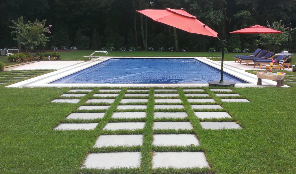 A backyard features a rectangular swimming pool with a stone walkway leading to it, surrounded by green grass, lounge chairs, and two large red umbrellas. Trees and shrubs line the background.