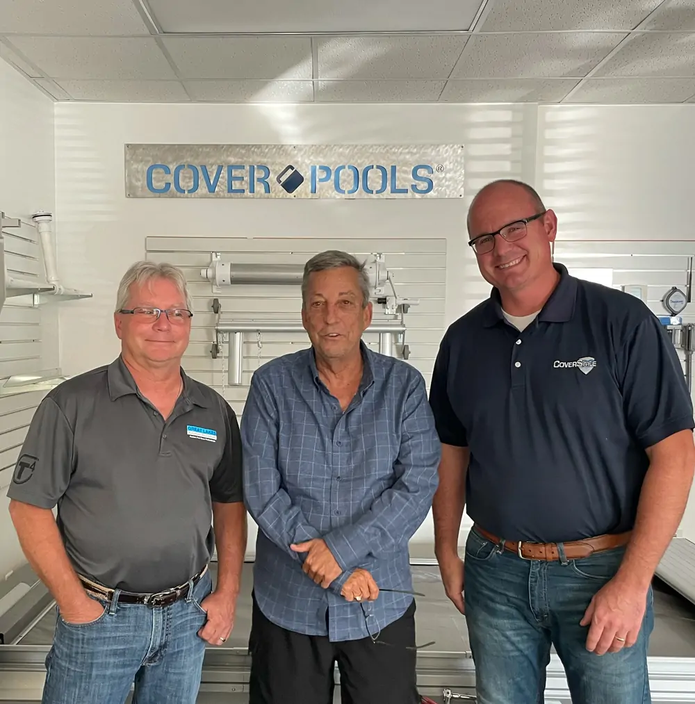 Three men stand smiling in front of a wall with a Cover Pools sign and pool equipment. The two men on the sides wear polo shirts and jeans, while the man in the middle wears a blue button-up shirt and slacks.