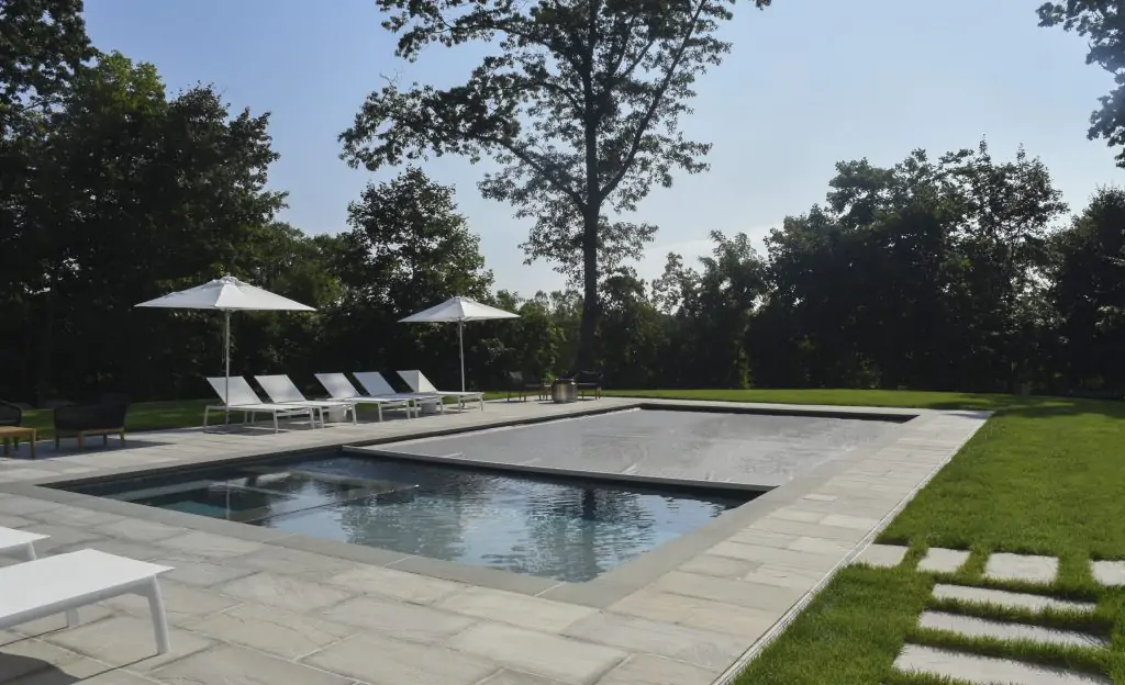 A modern backyard pool with a stone patio, surrounded by green grass and trees. Lounge chairs and white umbrellas are arranged on one side of the pool. The sky is clear and sunny.