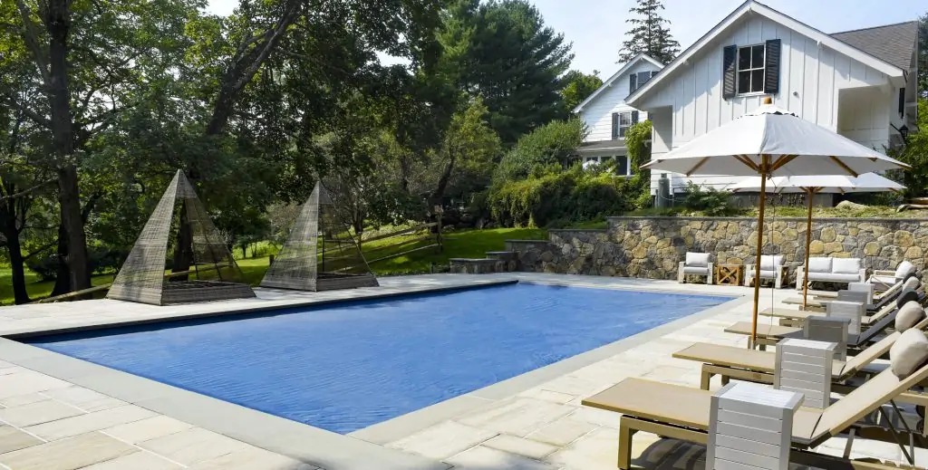 A rectangular outdoor swimming pool with lounge chairs and umbrellas beside it, surrounded by trees and a stone wall, with a white house in the background.