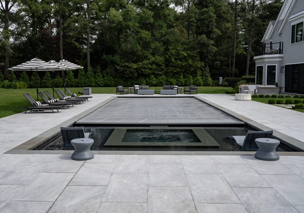 A modern backyard with a covered rectangular swimming pool, surrounded by lounge chairs with striped cushions, patio furniture, umbrellas, and a large gray house in the background, all set amid lush greenery.