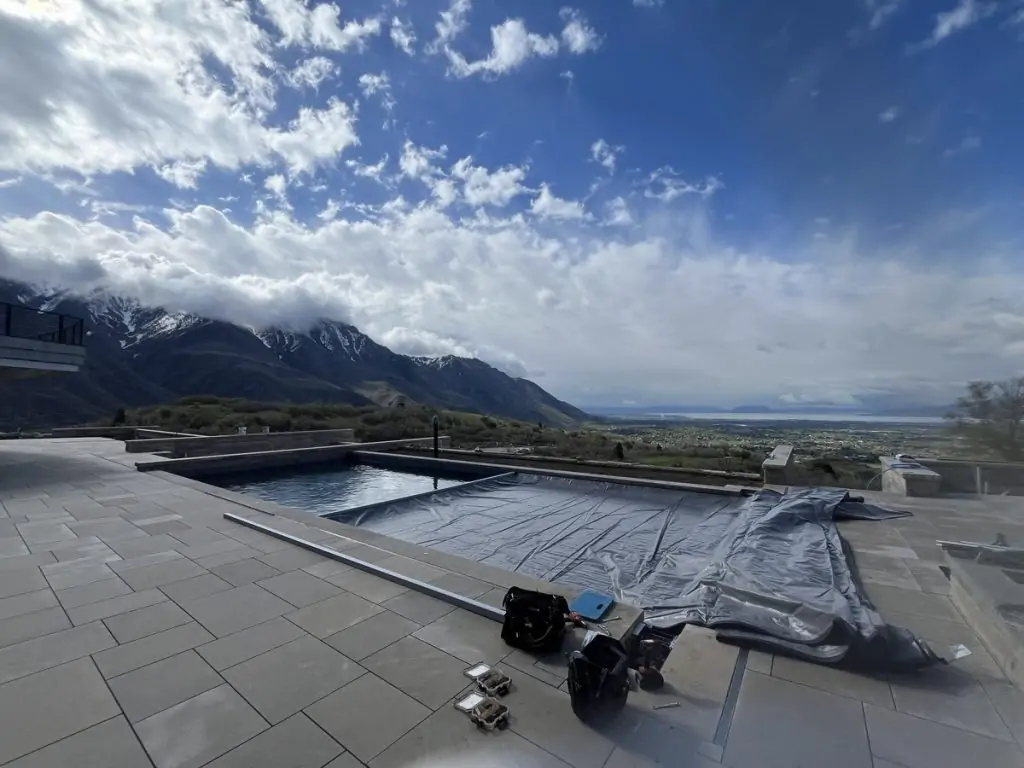 A modern pool with a partially open cover sits on a large stone patio overlooking a scenic valley and snow-capped mountains under a partly cloudy sky. Tools and bags are on the ground near the pool.