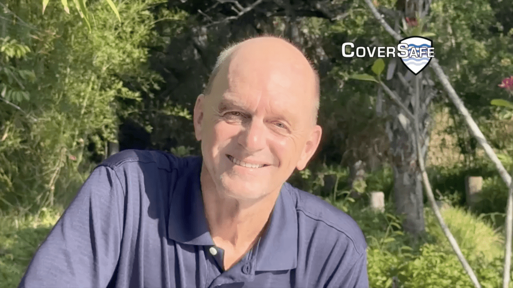 A smiling older man wearing a blue polo shirt sits outdoors with greenery in the background. The word CoverSafe with a shield logo appears in the upper right corner.