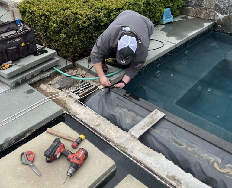 A person in a gray jacket and cap is kneeling by a pool, installing or repairing pool equipment with various tools, wires, and power drills on nearby stone slabs. A tool bag and garden hose are also visible.