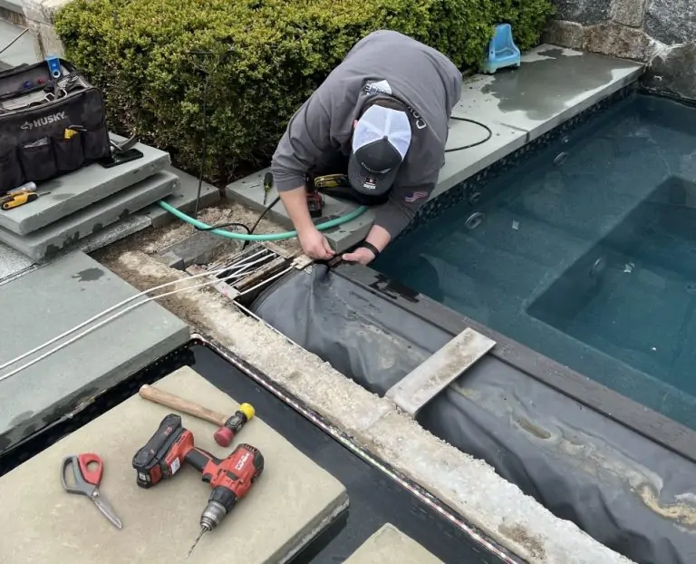 A person in a gray jacket and cap is kneeling by a pool, installing or repairing pool equipment with various tools, wires, and power drills on nearby stone slabs. A tool bag and garden hose are also visible.