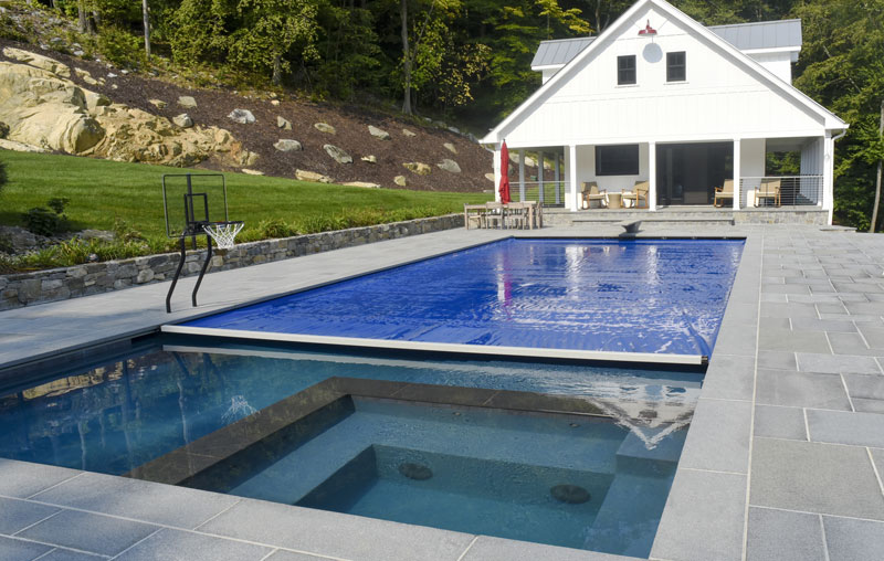 A modern backyard with a swimming pool partially covered by a blue pool cover, a built-in hot tub, a basketball hoop by the pool, and a white house with a porch in the background.