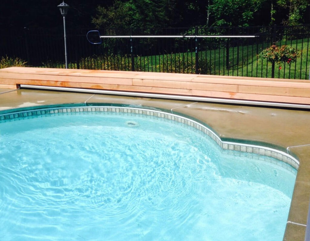 A backyard swimming pool with clear blue water is bordered by a curved concrete deck and a low wooden bench. A black metal fence and green lawn with bushes and flowers are visible in the background.
