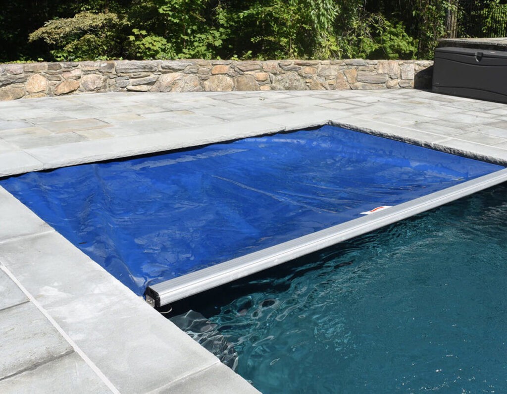 A rectangular pool with a partially closed blue safety cover, surrounded by a stone patio and a stone wall, with green foliage in the background.