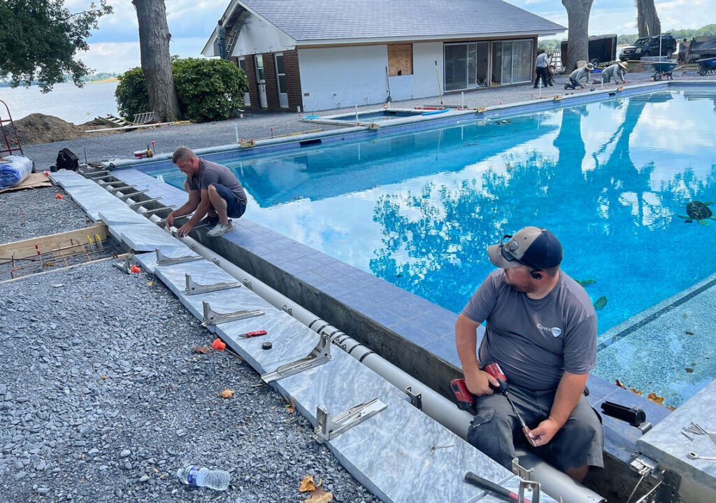 Two men work on the edge of an outdoor swimming pool, installing metal framework beside the water. Tools and construction materials are scattered around them. A house and trees are visible in the background.