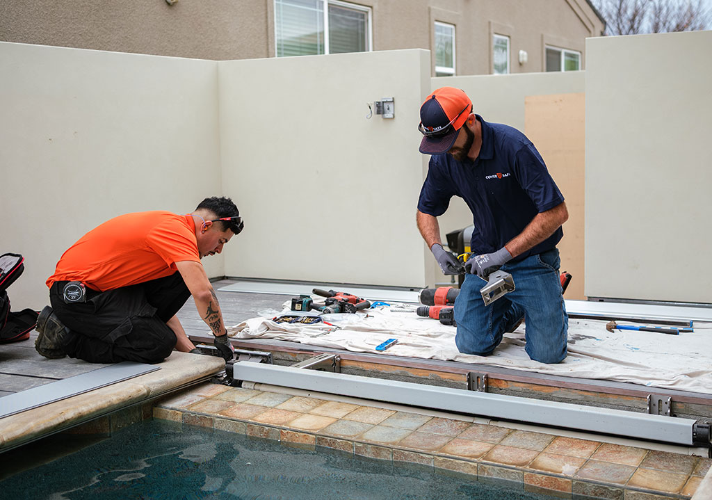 Two workers wearing safety gear and tools are installing or repairing a metal structure beside a swimming pool, with various tools and equipment spread out on a white cloth nearby.