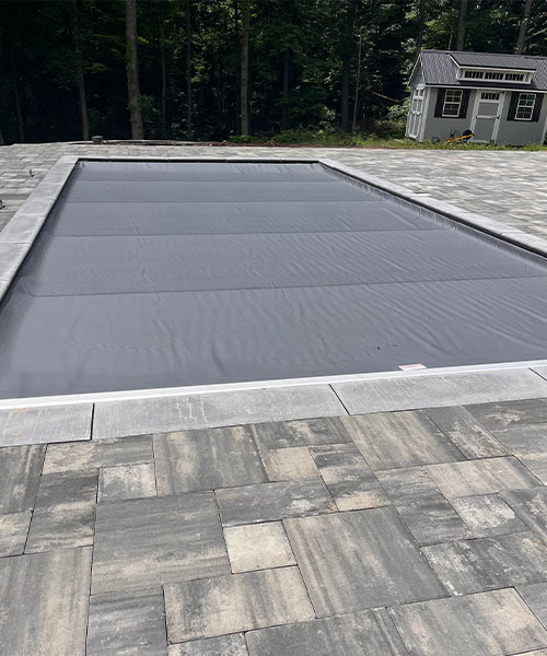 A rectangular swimming pool with a gray safety cover is surrounded by stone pavers. A small shed and trees are visible in the background.