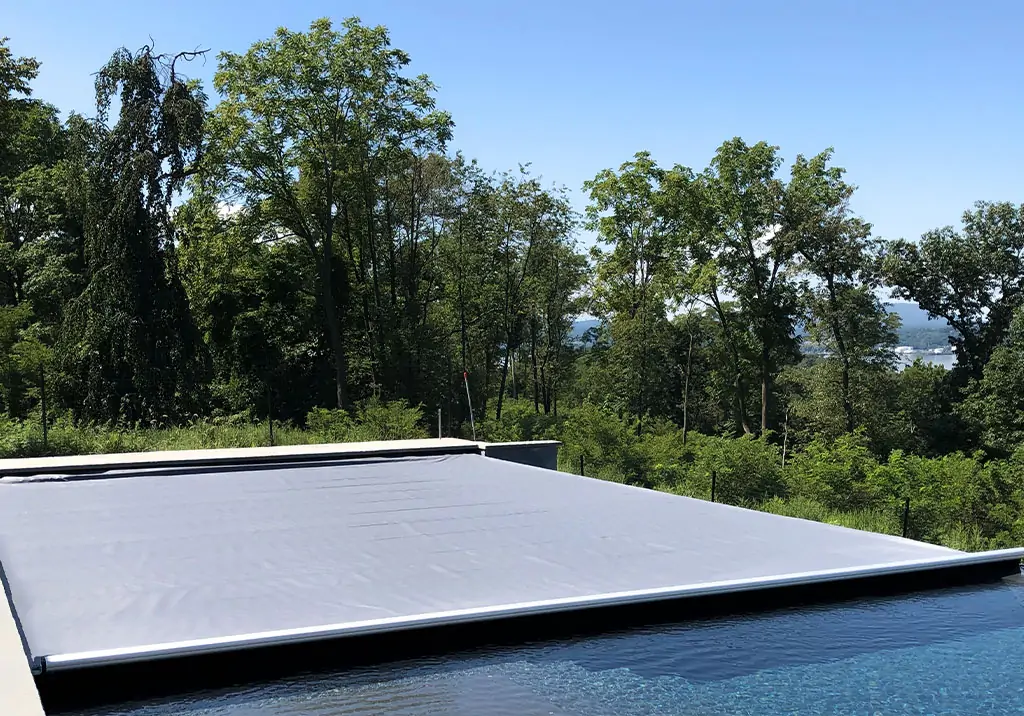 A swimming pool with a closed gray safety cover, surrounded by lush green trees and vegetation under a clear blue sky.
