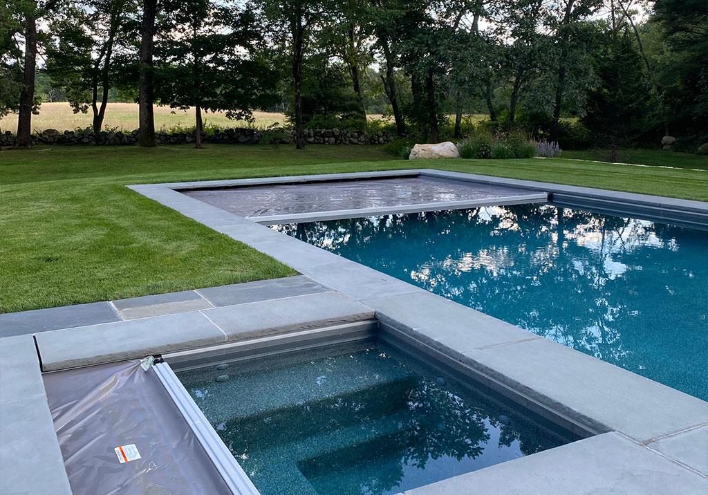 A modern backyard pool with a gray stone border features a separate hot tub area and an automatic pool cover partially extended. Green lawn and trees surround the pool.