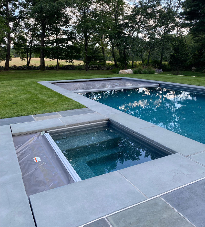 A modern backyard with a rectangular swimming pool and an adjacent hot tub, both surrounded by stone tiles and green grass, with trees and open space in the background.