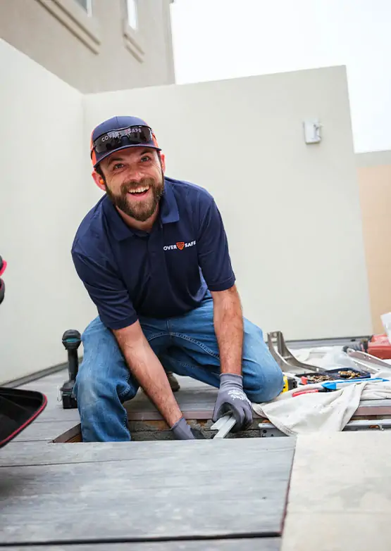 A smiling man in a navy polo shirt and cap kneels on the ground while installing or repairing equipment. Tools are spread nearby, and he is working outdoors near a building.