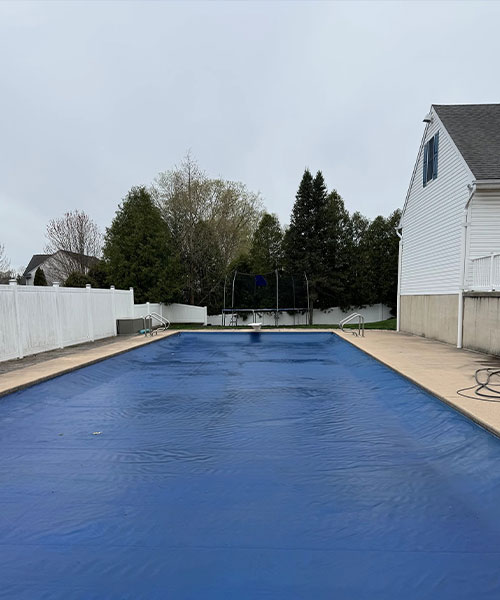 A rectangular outdoor swimming pool covered with a blue pool cover, surrounded by a concrete patio and white fence, with a house and trees visible in the background under an overcast sky.