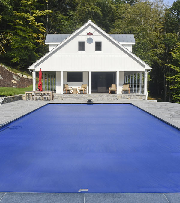 A white house with a porch sits behind a rectangular swimming pool covered with a blue pool cover. The area around the pool is paved, and there are trees in the background. Outdoor furniture is visible on the porch.