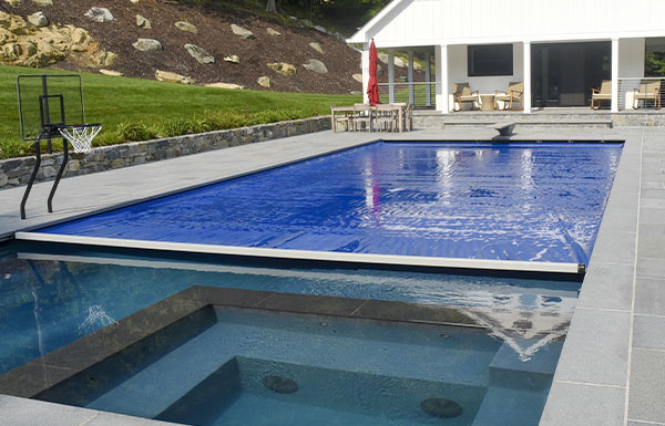 A backyard with a rectangular swimming pool partially covered by a blue pool cover, adjacent to a hot tub. There is a basketball hoop by the pool and a modern white house with outdoor seating in the background.