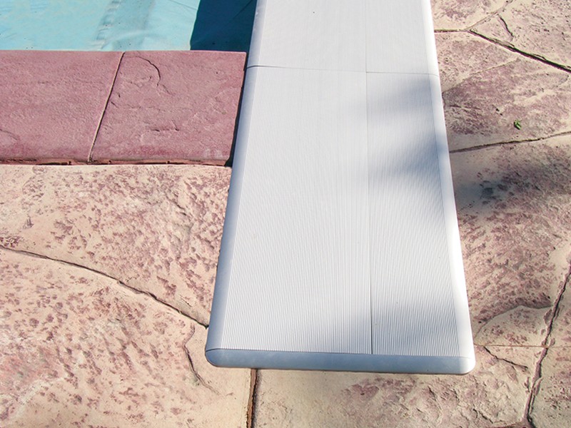 A close-up view of the end of a white diving board extending over a textured, reddish concrete pool deck, with part of a swimming pool visible in the upper left corner.