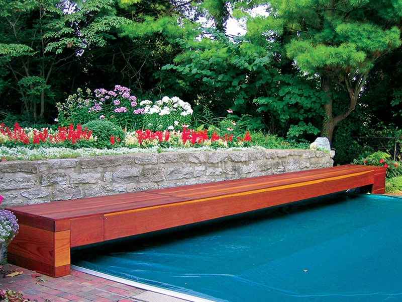 A long wooden bench sits next to a swimming pool covered by a blue pool cover, with a stone wall and colorful flowers and greenery in the background.
