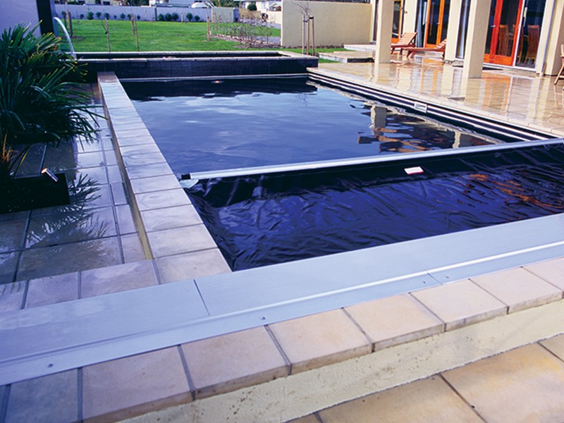 A modern rectangular swimming pool with a partially extended black safety cover, surrounded by tan tiles and a landscaped yard, next to a building with large glass doors.