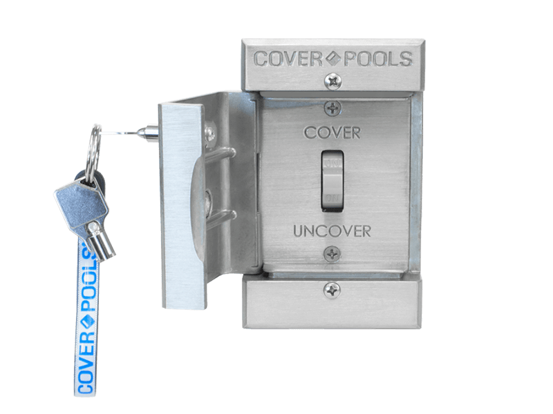 A metal wall-mounted pool cover control switch labeled COVER and UNCOVER, with a key inserted and a blue COVER POOLS tag attached to the keyring.