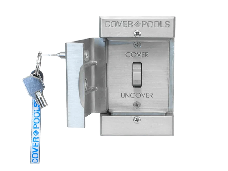 A metal wall-mounted pool cover control switch labeled COVER and UNCOVER, with a key inserted and a blue COVER POOLS tag attached to the keyring.