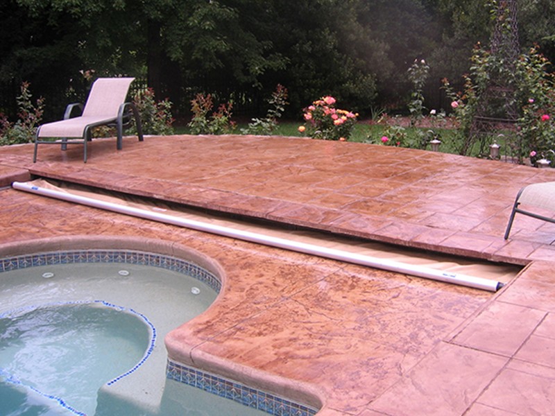 A retractable pool cover is partially extended over a circular pool and spa, surrounded by stamped concrete patio with lounge chairs and blooming garden in the background.