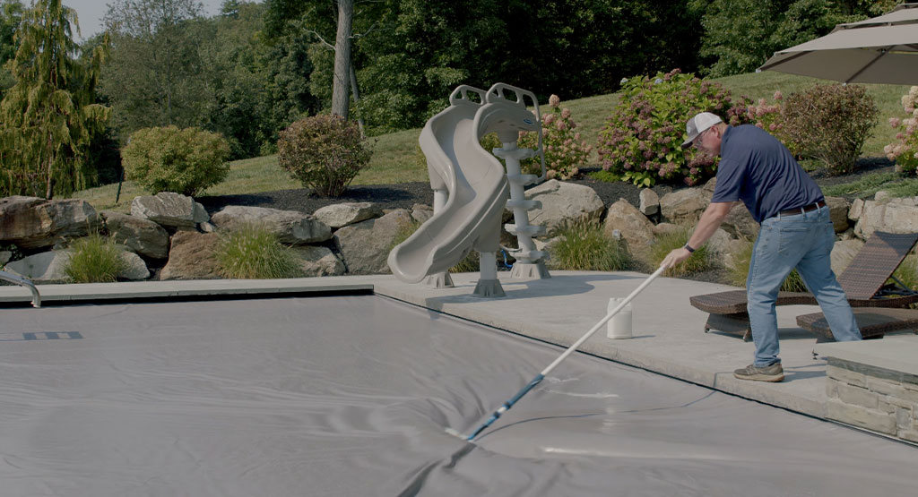 A man in a cap uses a long pole to clean or adjust a pool cover on an outdoor swimming pool, with a slide, lounge chairs, rocks, and greenery in the background.