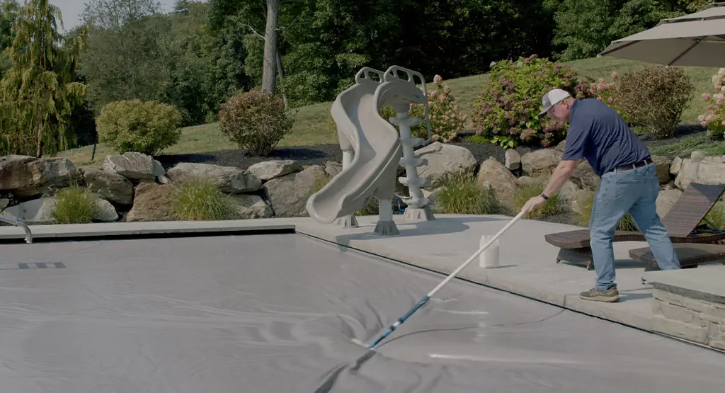 A man in a cap uses a long pole to clean or adjust a pool cover on an outdoor swimming pool, with a slide, lounge chairs, rocks, and greenery in the background.
