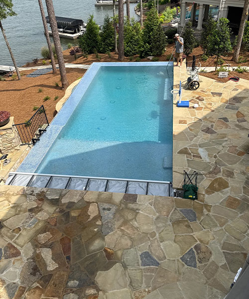 A rectangular swimming pool with clear blue water is surrounded by a stone patio. Trees and landscaping are in the background, and a person stands near the back corner by equipment next to a lake.