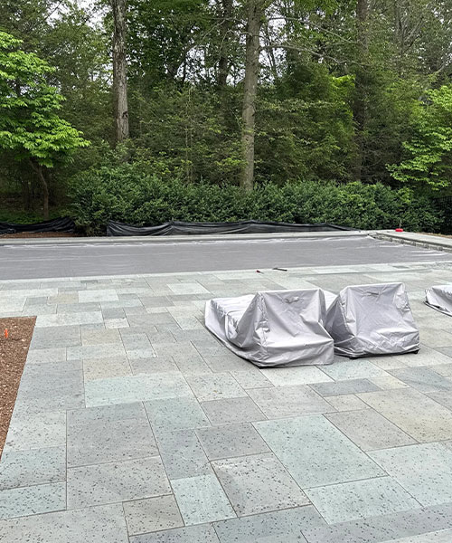 A stone patio with two pieces of outdoor furniture covered in gray tarps. In the background, there are trees, shrubs, and a paved area separated by a low barrier.
