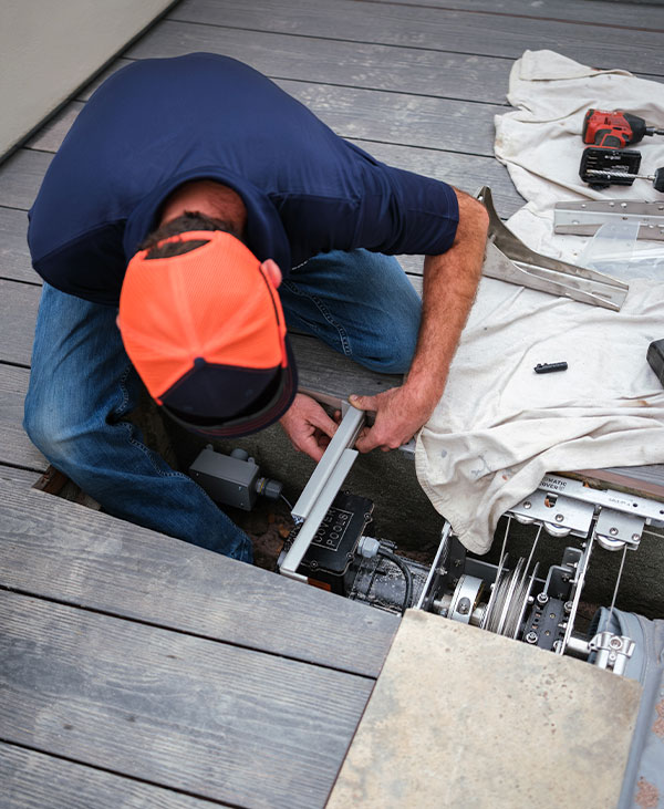 A person in a blue shirt and orange cap installs or repairs mechanical equipment beneath wooden deck boards, using tools and hardware spread nearby on a cloth.