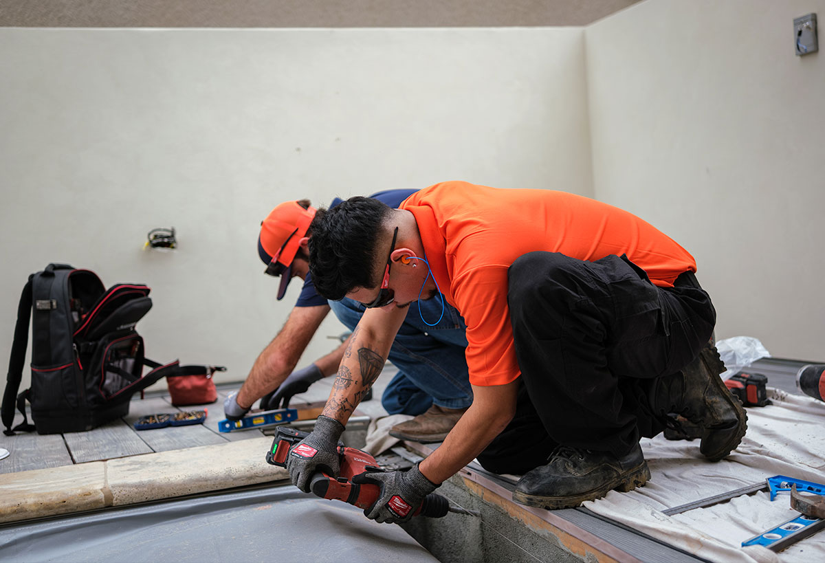 Two workers in orange shirts and black pants use power tools and measuring devices while installing or repairing tiles near a pool. Tools and a backpack are nearby, and the setting appears to be outdoors.