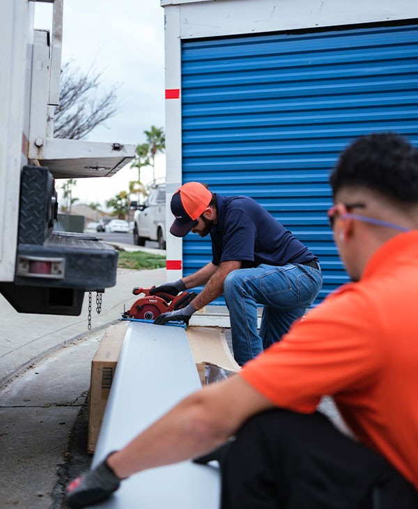 Two workers in orange shirts and caps cut a sheet of metal with a power saw on a sidewalk near a blue roll-up storage unit door. One operates the saw while the other observes nearby.