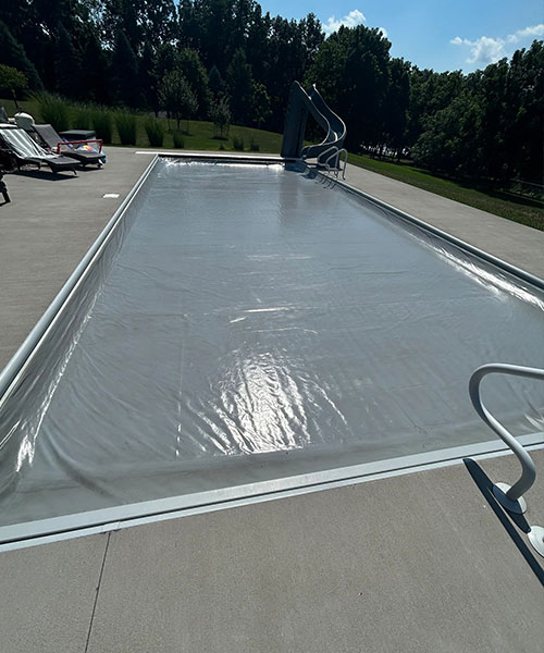 A rectangular outdoor swimming pool is covered with a silver safety cover. Lounge chairs and a table sit on the pool deck, and trees and grass surround the area under a sunny sky.