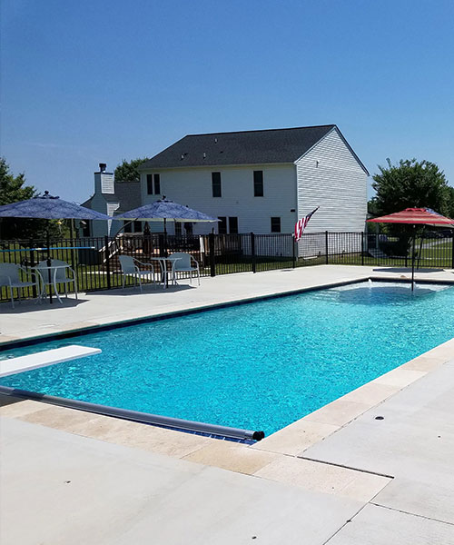 A clear blue swimming pool with white deck chairs, umbrellas, and tables around it, set in a fenced backyard with a two-story white house in the background under a clear blue sky.