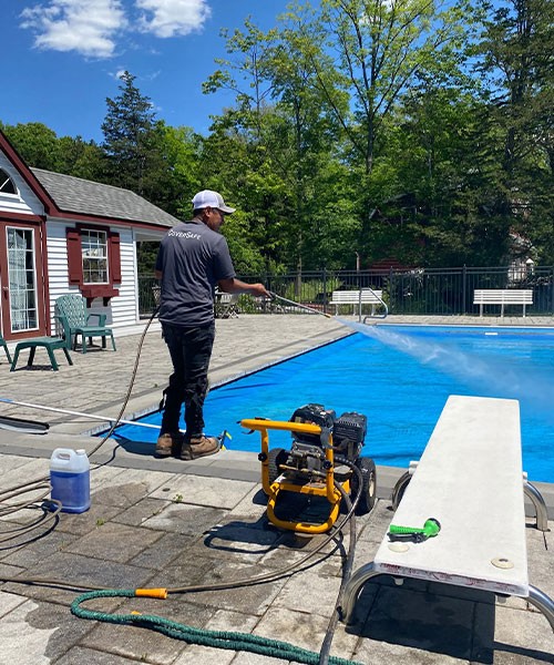 A person uses a pressure washer to clean the edge of an outdoor swimming pool, with cleaning equipment, a towel, and a bottle nearby on the poolside patio. Trees and a building are in the background.