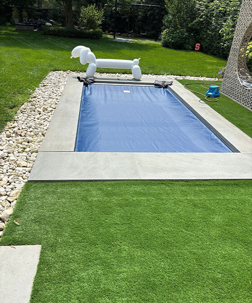 A small rectangular pool with a blue cover sits in a backyard with green grass, stone edging, and a large white balloon dog sculpture at one end. A blue toy and circular stone structure are nearby.