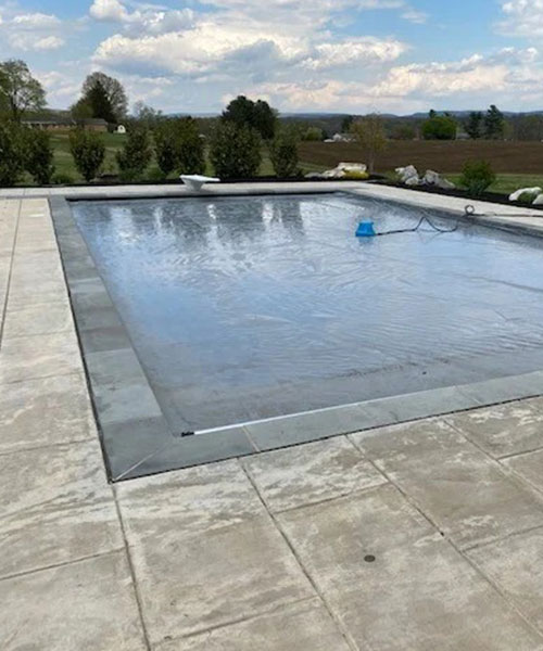 A swimming pool covered with a protective tarp sits in a backyard surrounded by a stone patio, with shrubs and trees lining the background under a partly cloudy sky.