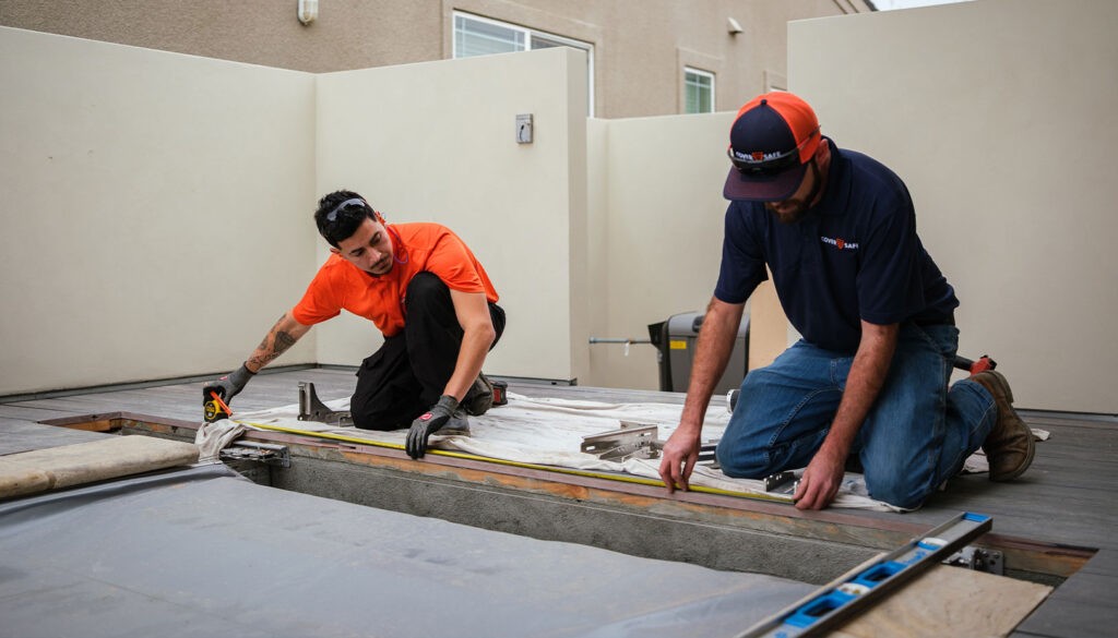 Two men kneel on a deck under construction, measuring a section with a tape measure and level. One wears an orange shirt and sunglasses; the other wears a navy shirt and cap. Tools and construction materials surround them.