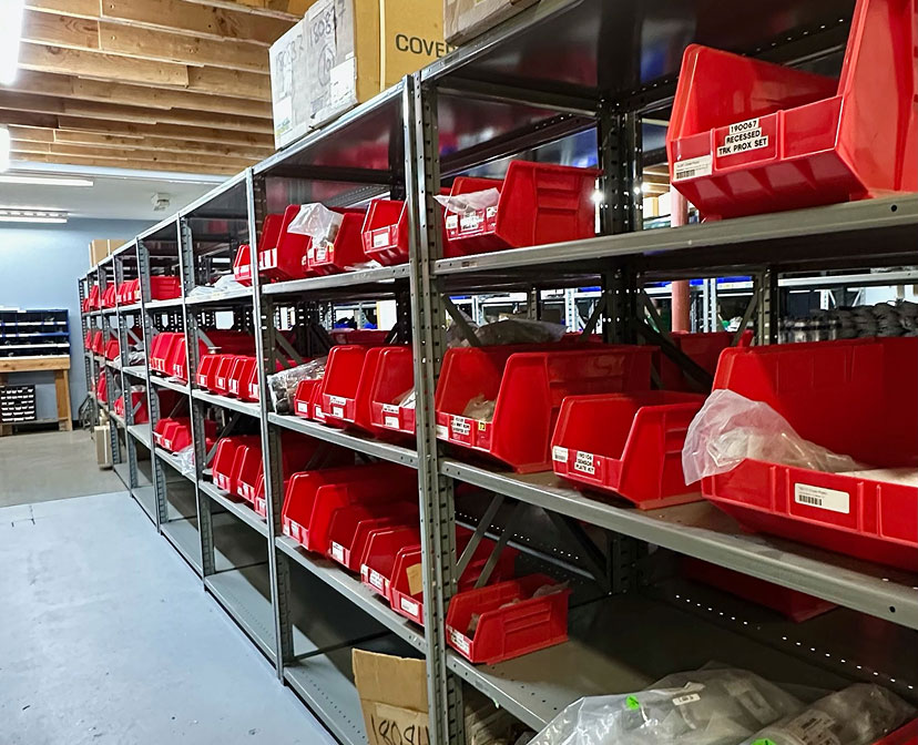 Metal shelves lined with red plastic bins, some containing small parts in plastic bags, are organized in a warehouse or storage room with fluorescent lighting and a concrete floor.